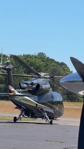 A helicopter with the registration number N6789R is parked on an asphalt surface, likely an airport tarmac. The helicopter is dark colored with a gold and black stripe along the side. Its rotors are spinning, and the tail rotor is also in motion. In the foreground, a blurred image of a propeller from another aircraft is visible on the right side of the frame. The background consists of a field with dry grass and a line of green trees under a clear blue sky. The lighting suggests it is daytime, likely sunny.