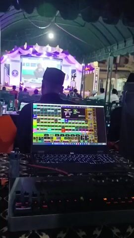 The video shows a dimly lit indoor setting, possibly an event tent, with multiple screens and equipment. In the foreground, a laptop displays a complex interface with many colored squares and buttons, likely for controlling video or audio production. A keyboard with illuminated keys is also visible in front of the laptop. To the right, a smaller mixing board with numerous illuminated knobs and sliders is present. Further back, several monitors are arranged in a row, showing various video feeds, some of which appear to be split into multiple smaller screens. One of these monitors displays a live video feed of people on a stage, with graphics superimposed. In the background, the outline of a stage with lighting and decorations is faintly visible. A few individuals are seated behind the equipment, seemingly operating the controls, with their backs to the camera. The overall impression is that of a live production control room, possibly for a broadcast or event.