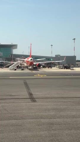 The video shows a view of an airport tarmac on a sunny day. In the foreground is an orange and white passenger airplane parked at a gate, with a set of stairs leading up to its door. Airport personnel are working around the plane. In the background, another plane is taking off. Further away, another plane, a Lufthansa aircraft, is visible on the ground. The tarmac is marked with yellow lines. The environment consists of airport buildings, lighting poles with red and white stripes, and various airport service vehicles.