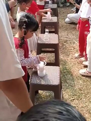 In an outdoor grassy area, a group of children are participating in what appears to be a game. Several children, mostly young girls wearing red and white, stand in a line facing away from the camera. They are positioned behind small brown stools, each with a clear plastic cup placed on top. The children are engaged in an action involving the cups, possibly pouring or transferring something. In the background, other people, including adults and children, are observing the activity. The environment suggests a festive or recreational event, possibly a school activity or a party, with the red and white attire possibly indicating a national celebration.