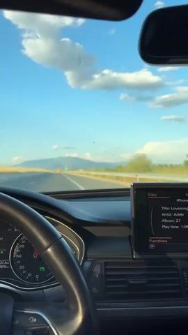The video shows a person driving a car on a highway on a sunny day. The dashboard displays the fuel consumption and the speedometer, which reads around 100 km/h. The car’s infotainment system is playing Adele’s “Lovesong.” The view from the windshield includes a blue sky with scattered clouds and distant mountains. The driver's hands are visible on the steering wheel.