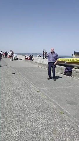 A man juggles with two balls on a promenade by a beach under a clear blue sky. He is wearing a patterned shirt and dark trousers. In the background, people are sitting on the pebble beach and walking along the promenade. There are several boats on trailers behind a low wall.