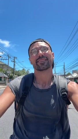 A man wearing glasses, a black baseball cap, and a grey t-shirt is riding on a motorcycle on a sunny day. He is looking at the camera and smiling. He has a backpack on. The road is paved, and there are buildings and utility poles on either side of the road. The sky is blue with a few white clouds.