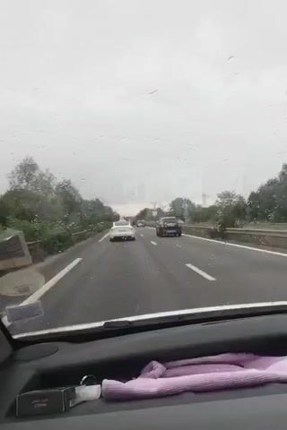 The media depicts a view from inside a car driving on a highway, likely in France, given the location context of Pomponne. The weather appears overcast and rainy, with water droplets visible on the windshield. The road is wet, and traffic is moderate.

The primary subjects are the cars on the highway: a white car and a dark SUV are prominent in the foreground, with other vehicles further ahead. The setting is a multi-lane highway surrounded by trees and vegetation. The time of day is unclear due to the overcast conditions, but it appears to be daytime.

On the dashboard of the car, several objects are visible. There is a black box, possibly for cigarettes or other small items, with white text on it that appears to read "LUMIERA" and possibly "CHATEAU". Next to it is a small white bottle, potentially hand sanitizer or a similar product. A folded purple cloth or towel is also on the dashboard.

The overall mood is neutral, conveying the experience of driving on a highway in rainy weather. There are no explicit indications of emotions, and the focus is on the journey and the environment. The road markings and signs (though not clearly legible) suggest a European highway system.