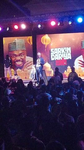 A low-angle shot of a stage presentation in a dark room filled with people. A man in a suit stands at center stage addressing the audience. Behind him, a large screen displays an image of a smiling man wearing a traditional cap. To the right of the screen, the words "SARKIN DARIYA" are displayed. Several figures stand or sit near the stage, including a cameraman. The audience, mostly dark silhouettes, fills the room. Spotlights of various colors illuminate the stage. The scene appears to be a comedy show or a similar live performance event.