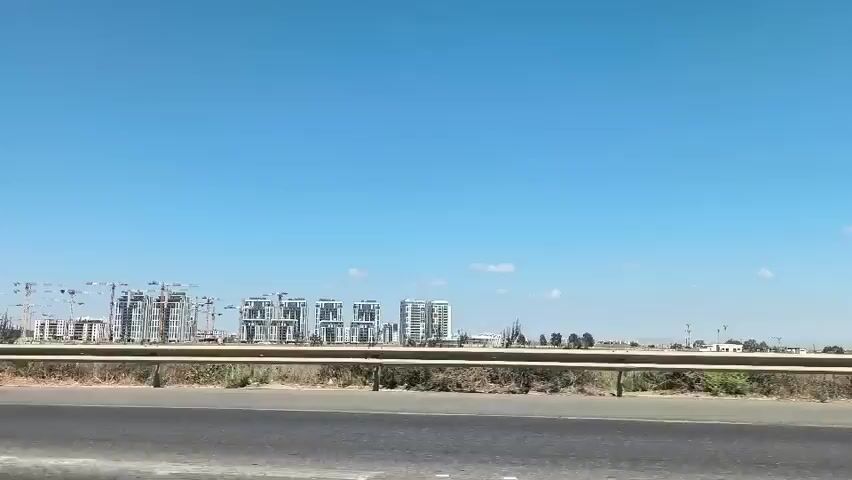 A black Mercedes-Benz GLE Coupe is driving on a road. The car is a dark color and has tinted windows. The driver, a man, is visible behind the steering wheel. The background shows a construction site with several tall buildings under construction and cranes. The sky is clear and blue.