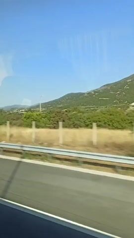 The video is a first-person perspective shot from inside a vehicle, looking out the side window. The vehicle appears to be on a highway. The scenery outside consists of a road, a metal guardrail, dry grass, green trees, and hills in the distance under a clear blue sky with some clouds. There are also signs visible, one with a red circle and a horizontal line, indicating a speed limit, and another sign with "80" in a red circle, also indicating a speed limit. A white car is visible ahead on the road. The sun is shining brightly. Text overlays in Ukrainian indicate "600 meters left."