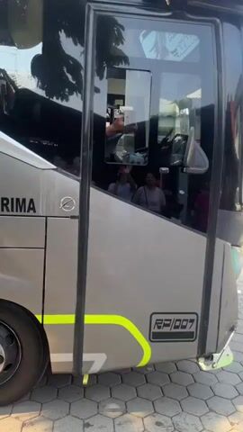 The video shows the side of a silver and gray tour bus named "Shela Alena". The bus has a dark tinted window with a person visible inside waving. The bus also has the text "PT. PUTRA ROSAN PRIMA" and a phone number "08563364610" written on its side. There are yellow and gray decorative stripes along the side of the bus. The bus is parked on a paved surface.