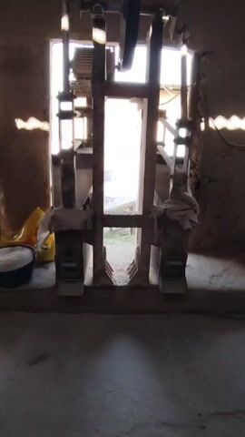 The camera, at floor level, shows a large wooden frame and machine in a semi-enclosed earthen-walled room, with the background light washing out visual detail. The machine appears to have an electric motor and belt drive at the top. The machine's frame is made of heavy timber and is set into a concrete floor. A yellow plastic bag and blue bucket with white contents sit to the left of the machine, on the floor. A bright opening in the back wall of the room, at the machine's center,  provides a view of a landscape. The machine is in operation, with components reciprocating, producing a rhythmic noise. The walls of the room are rough and uneven, made of earthen materials. The environment suggests a workshop or small industrial space in a rural setting.