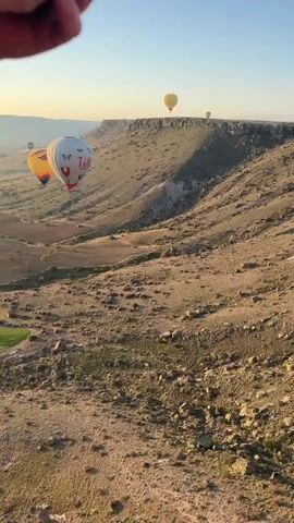 The video captures a scenic view from a hot air balloon, showcasing a rugged, arid landscape under a clear blue sky, likely during sunrise or sunset given the warm light and long shadows. Several hot air balloons are visible in the distance and mid-ground. One prominent white balloon clearly displays the text "Tally" along with other graphical elements. A yellow/orange balloon flies nearby, and two more, one yellow and one white, are seen farther away near a distinct cliff formation. The terrain below is characterized by dry, rocky ground with sparse patches of green and brown vegetation, sloping down towards a valley or canyon. The camera, seemingly from another hot air balloon, pans slowly to the right, offering a broader view of the expansive landscape and the distant cliff. The scene conveys a sense of adventure and tranquility, set in what appears to be a distinctive natural formation like those found in Cappadocia, Turkey, consistent with the provided location context of Yeşilhisar. The sound of a hot air balloon burner is audible at the beginning of the video.
