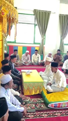The video shows a gathering of men sitting on a carpeted floor in a room. The room appears to be decorated with colorful banners on the wall and green curtains. In the center of the room, two young men in white clothing and caps are seated on cushions facing each other. In front of them is a low yellow table with items on it. Other men, also mostly wearing white or traditional clothing and caps, are seated around them in a semicircle. Some of the men are looking towards the two young men, while others are looking in different directions. One man on the right is holding a phone. The lighting suggests it is daytime.