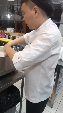 A person in a white chef's coat and a black chef's hat is working in a kitchen. They are standing next to a stainless steel counter and appear to be preparing food in a large metal pot. The person is using their hands to press down on something inside the pot, which is covered with what looks like parchment paper. In the background, there are shelves with various kitchen supplies and equipment. A black trash can is visible beneath the counter.