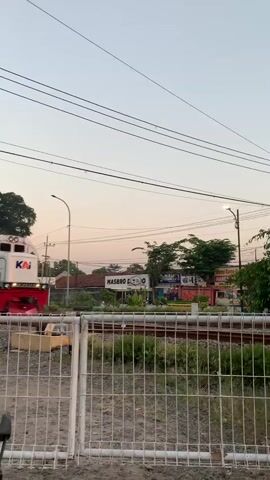 A white and red KAI train, marked "CC 203 98 01", moves from left to right across the frame. A person is visible in the train's cab. In the foreground, a white metal fence separates the viewer from the tracks. As the train passes, a railroad crossing signal with flashing red lights and a crossbuck becomes visible. Several individuals on motorcycles are stopped at the crossing, waiting for the train to clear. Power lines stretch across the sky, which has a light, possibly dusk or dawn, hue. Trees and buildings are visible in the background. A small red scooter or motorcycle is partially visible in the bottom left corner.