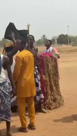 The video shows a group of men in an open, dusty field. Several are dressed in what appear to be traditional or ceremonial attire, including colorful wraps, beads, and a large, possibly ritualistic, costume made of rope-like material. One man carries a staff topped with a golden ornament. The attire varies, with some men in simple clothes and others in more elaborate garments. The men interact, gesturing and speaking, and one man plays a drum. In the background, there are buildings and trees, suggesting a town or city nearby. The lighting is bright, and the video appears to be shot outdoors on a sunny day.