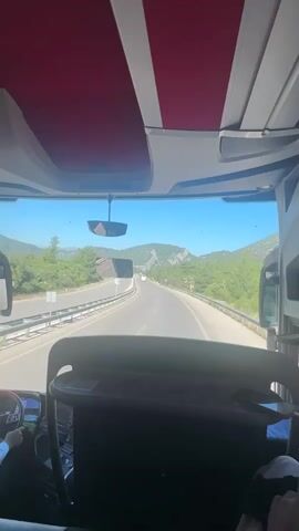 This is a first-person perspective video from inside a bus, looking forward through the windshield. The bus is traveling on a highway through a mountainous, green landscape. The sky is clear and blue, suggesting a sunny day. The time of day appears to be midday.

In the foreground, the interior of the bus is visible, including the driver's seat on the left, the dashboard, and the backs of seats in front of the viewer. The bus has a red and white interior ceiling lining.

On the road, other vehicles can be seen, including at least two other buses and a few cars. The highway has multiple lanes in each direction, separated by a barrier. The surrounding scenery consists of lush green trees and rocky mountains under a bright blue sky.

There is a speed limit sign visible on the right side of the road with a number and "km/h" indicating the speed limit. Another blue and white road sign is visible on the left side of the road. The overall impression is that of a scenic journey through a natural landscape. No specific emotions are evident, but the clear weather and open road suggest a pleasant travel experience.