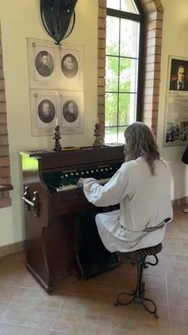 A person with long, light brown hair is seated on a stool playing an organ. The organ is made of dark wood and has white and black keys. The person is wearing a loose, light-colored robe with a patterned belt. Behind the organ, on the wall, are four framed portraits in a two-by-two grid. To the right of the organ is a tall window with a view of greenery. The room has tiled flooring and brick walls. A historical-looking poster is visible on the wall to the right.