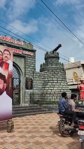 A vertical video shot from a moving vehicle shows a memorial site. The memorial features a stone structure resembling a fort gateway with an arched entrance and a cannon mounted on a turret. A statue of a man in traditional attire, holding a shield-like object, stands in front of the turret. In the foreground, three individuals are seated on a motorcycle, with the camera focused on the memorial. The sky is cloudy.