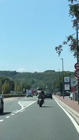 A motorcycle with two occupants travels down a paved road on a clear day. The road has multiple lanes and is bordered by sidewalks and trees. In the background, a forested hill is visible under a bright blue sky. Several cars are also present on the road. Signs for businesses such as Lidl and Assurances are visible along the roadside.