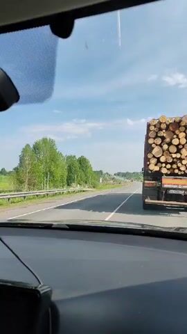 The video shows a road view from the inside of a car. The car is driving behind a large logging truck, fully loaded with logs. The sky is partly cloudy and there are trees to the side of the road. There is another smaller pile of logs on the side of the road further up ahead.