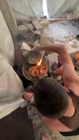 The video shows a person cooking food over an open fire in a makeshift shelter. The person, wearing a brown tank top and plaid shorts, is stirring a pot with a utensil. The fire is burning in a circular structure, likely a makeshift stove, and is producing flames and smoke. The surrounding environment appears to be a damaged or unfinished building, with concrete debris and white tarp visible. In the background, there are piles of debris, suggesting a location that has experienced destruction or is under construction. The lighting is natural, indicating it might be daytime. There are a few objects scattered around, including a plastic bottle and some bags. The scene suggests a basic, perhaps survivalist, cooking method in a challenging environment. No discernible text is visible in the video. The setting, with its debris and makeshift shelter, strongly implies a context of hardship or displacement.