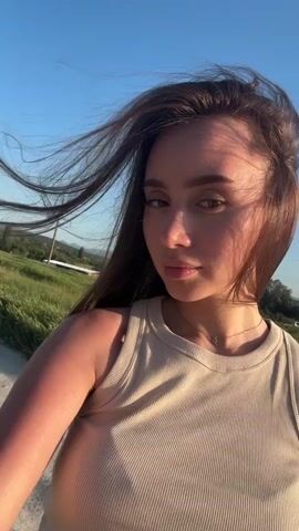 The video shows a young woman outside on a windy day. She has long, dark brown hair that is blowing in the wind, and she is wearing a light-colored tank top. The background includes a green field, trees, a blue sky, and other distant objects and vehicles. She looks at the camera and briefly touches her cheek. The sound of a fast-moving vehicle can be heard.