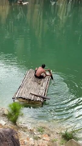 A medium, static shot shows a shirtless, dark-haired person on a makeshift bamboo raft in a quarry pond. The raft is positioned in the water near a rocky bank with a small evergreen plant. The pond is a milky green color with reflections of the quarry walls. The person is seated on the raft and is using a piece of wood as a paddle.