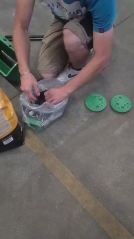 A person wearing khaki shorts and a blue t-shirt kneels on a concrete floor. In their hands, they hold a black wheel with a tread pattern, pulling it from a clear plastic bag. The bag also contains red and green items. To the right of the person, two green circular objects with dimples are on the floor. A yellow and black bag is visible on the left. A strip of brown tape runs diagonally across the floor.