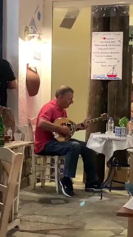 A group of people are enjoying themselves at an outdoor restaurant. A man plays a stringed instrument, possibly a bouzouki, while another man plays a guitar. Waiters bring food to tables. The atmosphere appears lively and festive, with music playing and people socializing. The restaurant has a rustic charm, with exposed wooden beams and traditional Greek decor. The lighting is warm and inviting, creating a cozy ambiance. The weather seems pleasant, as people are dining outdoors.
