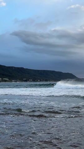 The video shows a surfer riding a wave in a coastal setting. The surfer, dressed in black, is positioned on the wave, skillfully balancing and moving along its face. The wave itself is moderate in size, with white foam cresting as it breaks. 

The background includes a stretch of coastline with a prominent mountain range covered in lush green vegetation. There are scattered buildings and structures along the coast, suggesting a small settlement or town. The sky above is partly cloudy, with a mix of blue sky visible amongst the dark, ominous clouds. The water in the foreground is choppy, with small waves lapping against the rocky shore.