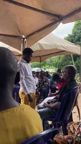 This media file captures an outdoor gathering under a tent, likely in Igbara, Nigeria. The scene is populated by a group of people, many seated on plastic chairs. In the foreground, the back of a person in a yellow shirt is visible. Towards the center, a man in a white shirt and yellow trousers is standing and interacting with two individuals seated on blue chairs. One of these individuals, in a red shirt, is holding a document and appears to be writing, while the other, in a black shirt, looks on. The man in the white shirt gestures towards the document.

In the background, more people are seated, and a building with red walls and a white roof is visible. The setting appears to be a community event or meeting, possibly related to some form of registration or administrative process given the presence of documents and writing. The atmosphere seems engaged, with individuals focused on the task at hand. The lighting suggests it is daytime, likely mid-morning or afternoon, with overcast skies hinting at possibly humid or rainy weather typical of tropical regions. The presence of trees and lush greenery in the background further reinforces the outdoor, possibly rural or semi-urban, setting. Some visible text on a blue chair in the background reads "N DU 1201720".