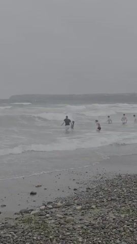 The video shows a group of people playing in the ocean on a cloudy day at Tramore, Ireland. The waves are moderate and there appear to be several individuals in the water, some closer to shore and others further out. The scene is set on a pebble beach with some sparse vegetation in the foreground. The sky is overcast, suggesting it is either early morning, late afternoon, or simply a day with no sun. The overall mood is relaxed and recreational, with people enjoying the water despite the weather conditions. There is no visible text in the media file.