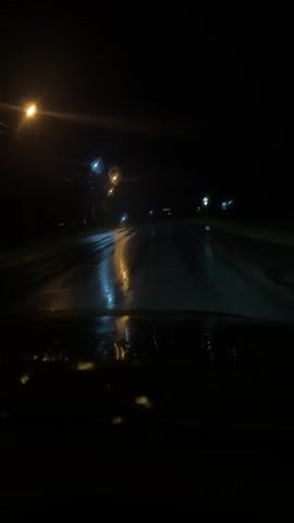 The video shows a view from inside a vehicle driving on a wet road at night. Streetlights and distant lights are visible, reflecting off the wet asphalt. The road appears to be empty of other vehicles. The windshield of the car is also wet, with streaks from water.