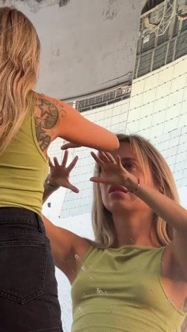 Two young women are in front of a large, curved, reflective surface, possibly a mirrored sculpture or building. One woman, seen from the back and left, has long blonde hair and is wearing a lime green tank top and black denim shorts. She has a tattoo on her left shoulder blade with stars and other designs. She is reaching out with her left hand towards the other woman. The second woman, seen from the front and right, also has long blonde hair and is wearing a lime green tank top. She is facing the reflective surface, and her hands are near her face, one hand covering her eyes and the other near her temple. There are water droplets on the reflective surface, and the background shows a distorted reflection of a building with a grid-like structure. The lighting suggests it is daytime.