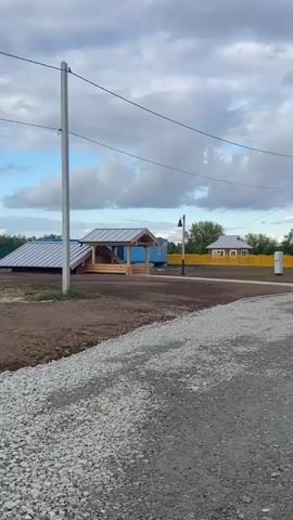 The video pans across a rural landscape under a cloudy sky. In the foreground, there is a gravel path. Behind it, a dirt area transitions to a grassy area with freshly planted sod and some construction materials. Several wooden houses with yellow fences are visible in the background. A wooden gazebo with a metal roof and blue accents is in the mid-ground, near a lamppost. Further down the road, more houses and trees can be seen. The scene appears to be a village or a newly developed residential area.