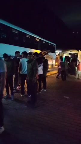 A group of people stand in front of a large white bus at night. The bus has its doors open and lights on, illuminating the area. Some individuals are gathered around the entrance of the bus, while others stand further back. Luggage, including backpacks and duffel bags, is visible on the ground near the bus. The scene appears to be a bus station or a similar transit area, with patterned paving stones on the ground and building structures in the background. The lighting is dim, with the bus's headlights and interior lights being the primary sources of illumination. The people are dressed in casual attire, and some appear to be interacting with each other or preparing to board the bus. The overall atmosphere suggests a moment of transition, possibly during a stopover or at the beginning/end of a journey.