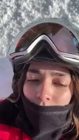 A person wearing a red winter coat, white ski helmet with goggles, and a black neck warmer is lying in the snow. The person extends one arm out to the side, and skis are visible next to them. The video is taken from above, capturing the person's face and upper body against the white snow. The weather appears sunny.
