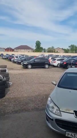 The video shows a large parking lot filled with cars, predominantly sedans and SUVs of various colors, including silver, black, white, and red. In the foreground is a silver Peugeot car with a visible license plate. The parking lot surface is gravel and asphalt. Several buildings with orange roofs are visible in the background, along with trees and a light pole. The sky is blue with scattered clouds. The camera seems to be held by someone standing near the Peugeot. The camera pans to the right, revealing more cars in the lot.