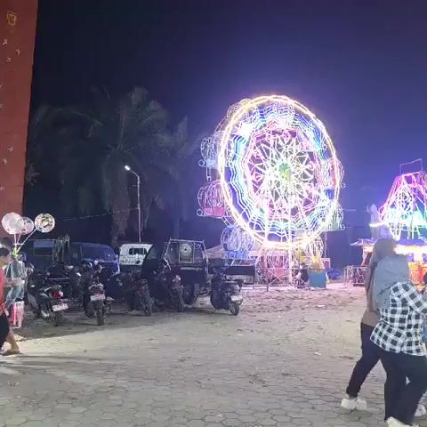The video captures a nighttime scene at what appears to be a fairground or amusement park. A large Ferris wheel, adorned with colorful, bright lights, dominates the center of the frame. To the right of the Ferris wheel, another amusement ride, possibly a drop tower or a similar structure, is visible, also lit up with vibrant lights. In the foreground, several people are present. On the left, two individuals are walking away from the camera. One person is wearing an orange t-shirt and black pants, while the other is wearing a white t-shirt and dark pants. A group of motorcycles is parked on the left side of the scene. In the mid-ground, several vehicles, including a white truck and a dark-colored van, are parked. The ground is paved with what looks like cobblestones or interlocking bricks. The overall environment suggests an outdoor event at night, characterized by artificial lighting from the rides and possibly other attractions. The sky is dark, indicating it is well after sunset. There are also palm trees visible in the background on the left, suggesting a tropical or subtropical location. Some of the people in the foreground appear to be observing the rides, while others are walking through the area. The scene conveys a lively and festive atmosphere.