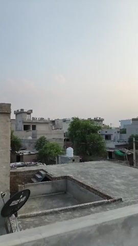 The video is a vertical, panning shot of a rooftop view of a residential area with multiple buildings and trees under a hazy sky. In the foreground, a concrete rooftop is visible with some debris. To the left, a satellite dish is mounted on a metal pole. Beyond the foreground rooftop, several buildings of varying heights and architectural styles are seen. Some buildings are grey, while others are lighter in color. Trees with green foliage are interspersed between the buildings. There are also signs of daily life, such as clothes hanging to dry on a clothesline between two structures, and satellite dishes mounted on some rooftops. A water tank is visible on one of the rooftops. The overall impression is of a moderately populated urban or semi-urban environment. The lighting suggests it is either early morning or late afternoon due to the soft, diffused light.