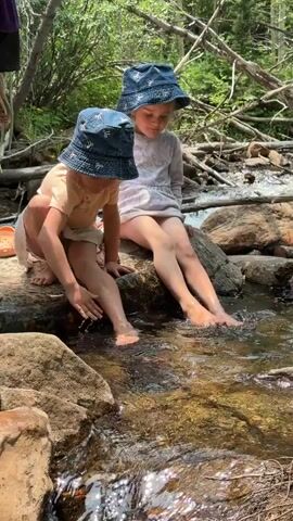 This media file shows two young children sitting on rocks at the edge of a stream. Both children are wearing blue bucket hats with a white pattern. The child on the left is wearing a light-colored t-shirt and shorts, with their left leg dangling in the water. The child on the right is wearing a light-colored long-sleeved shirt and appears to be wearing shorts as well. Their legs are bent, and their feet are also near or in the water. The stream is shallow and clear, with sunlight reflecting off the surface. Large rocks and tree branches are visible around the stream, with lush green foliage in the background, suggesting a wooded natural setting. The overall atmosphere appears peaceful and leisurely, with the children enjoying the cool water on a warm day. The lighting suggests it is daytime, likely late morning or early afternoon, with bright sunlight filtering through the trees. No visible text is present in the media file.