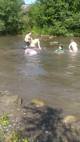 In this media file, a group of people are enjoying a day at a river. The scene is set in a natural environment with lush green trees bordering the water. The river itself appears to be shallow and somewhat murky, with a rocky bed visible along the shore.

The subjects are primarily children and possibly a few adults, all dressed in swimwear. They are engaged in various water activities. One person is splashing vigorously, creating a spray of water. Others are wading or swimming in the river. The overall atmosphere seems joyful and relaxed, suggesting a casual outing on a warm day.

The time of day appears to be daytime, likely late morning or early afternoon, given the bright sunlight casting shadows on the riverbank and the overall illumination of the scene. The weather seems pleasant and conducive to outdoor water activities. The location context of Hoverla, Ukraine, suggests a rural or mountainous setting, which aligns with the natural landscape depicted.

There are no visible signs or text within the frame of this media file. The focus is entirely on the people and their interaction with the natural environment. The image captures a moment of simple summer fun and recreation.