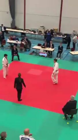 In an indoor venue, a judo match concludes with the participants bowing on a mat of red over green. A female referee in a black suit stands between them. Spectators, including officials seated at tables, applaud. The video then cuts to a presentation ceremony where a man in a black shirt and jeans places a medal around the neck of a female judoka standing on the top tier of a podium. Banners bearing the name "Orpi" stand behind them. A climbing wall with various grips is visible in the background. People in judo gis and civilian clothing observe the event.