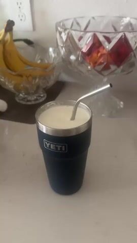 The video shows a dark blue YETI cup filled with a light-colored liquid, possibly a smoothie or milkshake, with a silver metal straw inserted. The cup is on a white countertop. In the background, there is a clear crystal bowl containing bananas, and another crystal bowl with red fruits, possibly apples or peaches. A power outlet is visible on the wall in the upper left corner. The lighting is natural, suggesting the scene is indoors during the day.