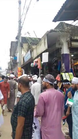 Here's a forensic description of the video:

**Setting:** The scene takes place on a crowded street in what appears to be a densely populated urban area in South Asia. There are multi-story buildings lining the street, many with shops on the ground floor. Electrical wires crisscross overhead, obscuring the sky.

**People:** A large number of men are present, many of whom appear to be wearing traditional Muslim clothing. Some have white skullcaps and long robes. A few individuals can be seen with headbands, some displaying flags or other symbols.

**Objects:** Visible objects include:
*   Buildings of varying heights and conditions, some with signage in a local language.
*   Electrical poles and a dense network of wires.
*   Street vendors selling goods from carts and stalls.
*   Signs and banners in the local language.

**Actions:** The majority of the people are walking down the street. The group appears to be a procession or gathering, possibly a religious or cultural event.

**Interactions:** The men are walking alongside each other, some conversing. There doesn't seem to be any aggressive or violent interaction visible. The crowd seems to be generally moving in the same direction.

**Environment:** The atmosphere appears to be busy and potentially noisy, with the sounds of people talking and moving. The lighting suggests it is daytime.