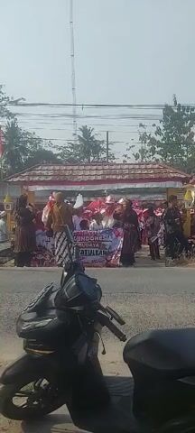 A group of people are participating in a parade or procession. In the foreground, a dark-colored motorcycle is parked. In the background, there is a building with a red-tiled roof and white trim. A banner is displayed with text that is partially visible, showing "ANANTI INDONESIA" and "BUDAYA SATULLAH". Several individuals are dressed in traditional attire, including headscarves and patterned clothing. Some are holding flags, and one person is standing on an elevated platform, holding up a white object. The overall atmosphere suggests a cultural or celebratory event. The sky is bright and clear, indicating a daytime scene.