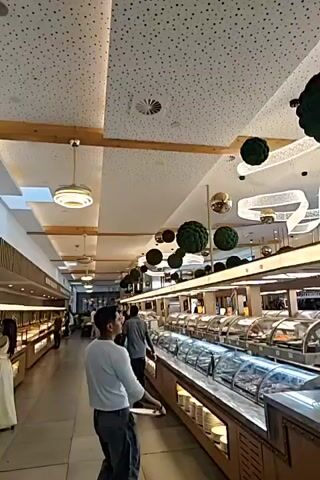 This media file captures the interior of a buffet-style restaurant. The scene is well-lit and appears to be during the daytime, though no windows or external views are clearly visible to confirm the weather. The setting is modern and decorative, with a textured ceiling adorned with small dark dots, interspersed with wooden beams. Various spherical decorations, some appearing to be clusters of green fabric resembling moss balls and others metallic gold, are suspended from the ceiling. Contemporary light fixtures, including a circular pendant light and ribbon-like illuminated structures, add to the ambiance.

The primary subject of the footage is a long buffet line filled with various food containers, mostly covered by glass to maintain freshness and temperature. These containers appear to hold a wide array of dishes, suggesting a diverse menu. In the foreground, a man in a white shirt and dark pants is walking along the buffet, holding a white plate, indicating he is in the process of selecting food. Other individuals can be seen further down the buffet line and in the background, suggesting the restaurant is open and patrons are dining.

A prominent object in the foreground is a Pepsi-branded soda fountain machine, indicating a beverage service is available. The machine has multiple spigots and bottles of Pepsi. Behind the buffet, there are glimpses of additional food displays and seating areas with people. The overall atmosphere appears relaxed and casual, as is typical of a buffet dining experience. There is no visible text within the frame, other than the Pepsi logo on the soda machine. The location context provided, Sint-Pieters-Leeuw, Belgium, helps to place this scene within a specific geographical area.