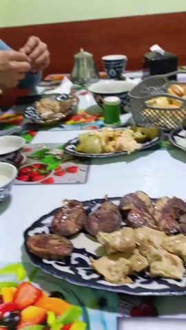 The camera focuses on a table laden with food. In the foreground, a plate displays cooked meats and doughy dumplings. Behind it, another plate holds pickled vegetables and what appears to be onions. Two traditional teacups with blue patterns are placed towards the left, and a metal basket filled with fried food sits on the right. In the background, a person with a blue sweater is visible, manipulating a piece of tissue or napkin, seemingly preparing for a meal. A silver teapot and various containers are also present on the table. The tablecloth features a colorful, fruit-themed design, with strawberries and other fruits visible. Overall, the scene suggests a meal setting with a variety of dishes ready to be enjoyed.
