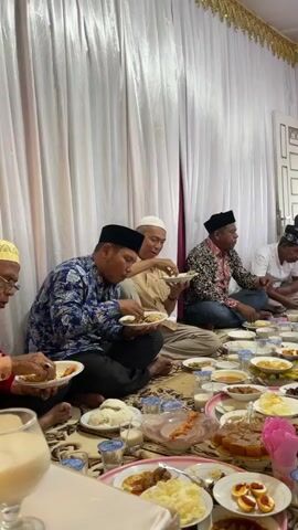 The video captures a communal meal, likely a religious gathering or celebration, taking place indoors in Teungoh Baro, Indonesia. A group of men, dressed in traditional attire including peci (caps) and long-sleeved shirts, are seated on the floor around a large spread of food. The room is decorated with white curtains and gold trim, suggesting a formal or festive occasion.

The food is presented on numerous platters and plates, arranged on a patterned rug that covers the floor. Dishes include rice, various curries or stews, fried items, and what appear to be hard-boiled eggs. Drinks, possibly milk or water, are also visible. The people are actively eating, some are engaged in conversation, and their expressions suggest a sense of community and shared enjoyment. The lighting appears to be natural, coming from an open doorway or window in the background, indicating it is likely daytime. The atmosphere seems warm and communal, indicative of a cultural or religious event common in Indonesia.