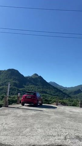 The video shows a view of a residential area with mountainous landscape in the background. The camera moves along a paved road, showing houses and garages on either side. The mountains are covered in trees, and the sky is clear and blue. A few cars are parked in front of houses or under carports. Overhead, there are electrical wires running alongside the road.