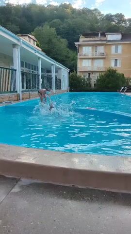 The video shows two children playing in a swimming pool. One child is wearing red swim trunks and is splashing around in the water. The other child, wearing a mask and what appears to be a blue swimsuit, is standing at the edge of the pool and appears to be observing. The pool is surrounded by a concrete deck, and in the background, there are buildings and lush green trees. The sky above is partly cloudy.
